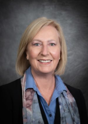 A woman with blonde hair smiles at the camera, wearing a blue collared shirt, patterned scarf, and dark jacket against a gray studio background.