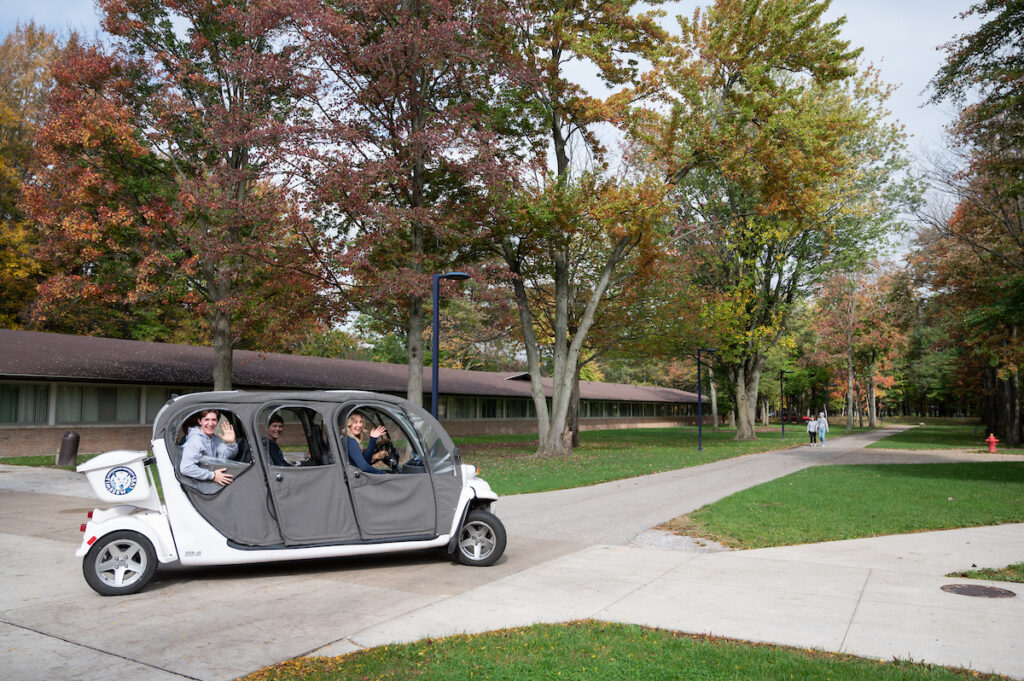 A small white electric shuttle with four passengers drives on a sidewalk through a tree-lined area on a partly cloudy day, offering a convenient way to visit campus.
