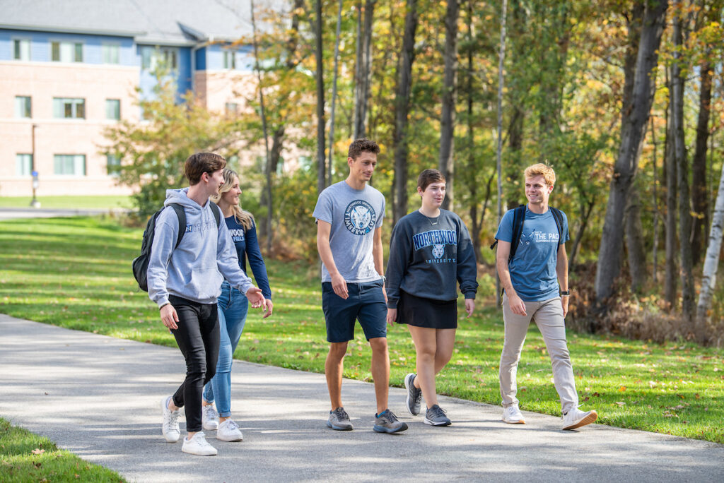 Five students walk together on a campus path lined with trees, wearing casual clothing and university-branded apparel—capturing the spirit of a Visit Campus experience.