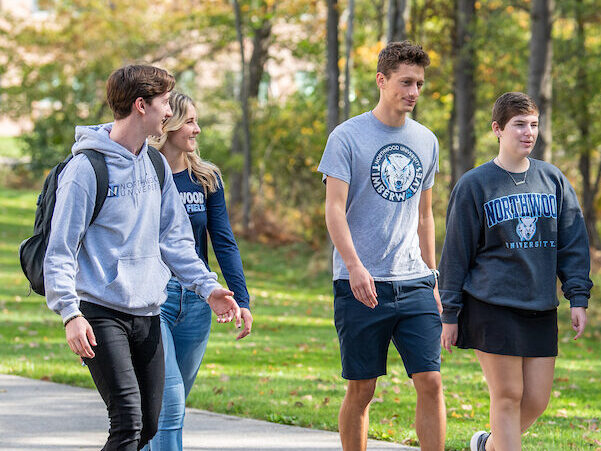 Five students walk together on a campus path lined with trees, wearing casual clothing and university-branded apparel—capturing the spirit of a Visit Campus experience.
