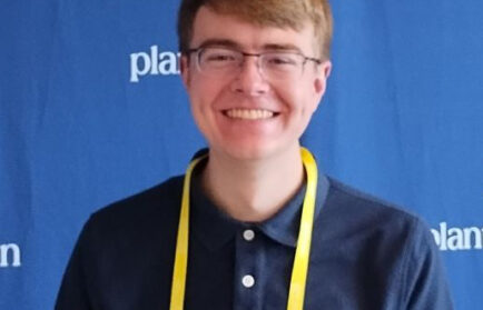 A person wearing glasses and a dark polo shirt with a yellow lanyard stands in front of a blue backdrop with partial white text, possibly attending an event focused on nuclear safety.