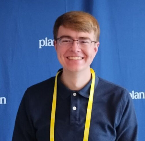 A person wearing glasses and a dark polo shirt with a yellow lanyard stands in front of a blue backdrop with partial white text, possibly attending an event focused on nuclear safety.