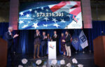 A group of people stand on stage applauding, with a large screen behind them displaying "$73,215,301" for the When We Are Free Campaign over an American flag graphic. Various flags are visible to the right.
