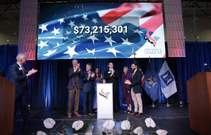 A group of people stand on stage applauding, with a large screen behind them displaying "$73,215,301" for the When We Are Free Campaign over an American flag graphic. Various flags are visible to the right.