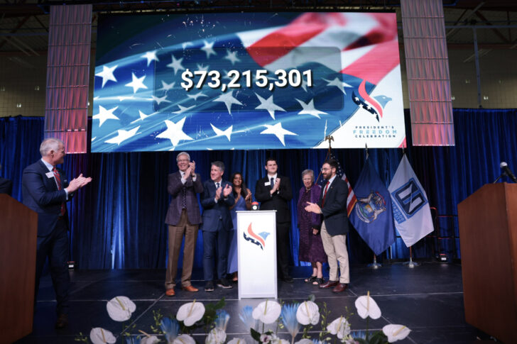 A group of people stand on stage applauding, with a large screen behind them displaying "$73,215,301" for the When We Are Free Campaign over an American flag graphic. Various flags are visible to the right.
