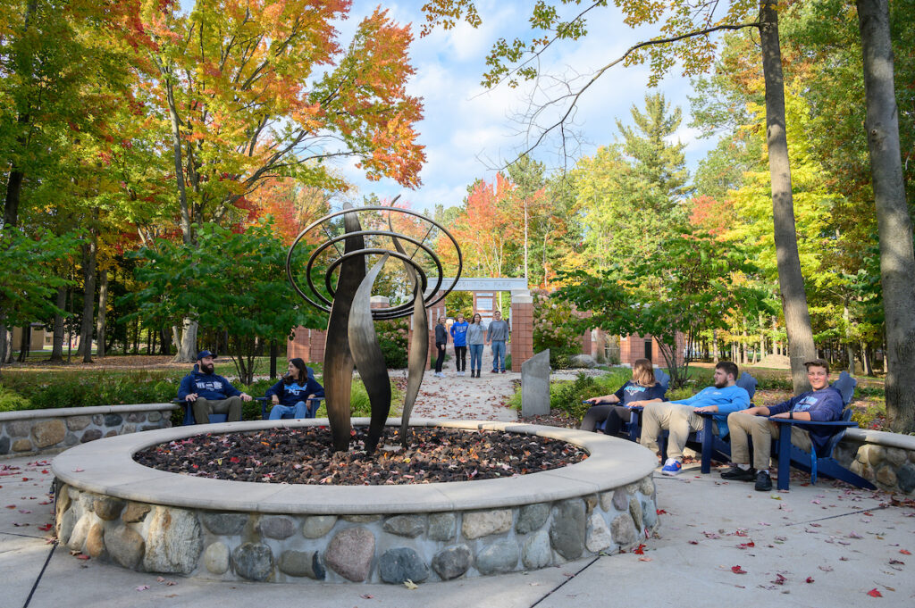 People sit and walk in a tree-filled outdoor plaza with a modern abstract sculpture in the center, surrounded by a stone wall and autumn foliage. Visit campus to experience this vibrant space firsthand.