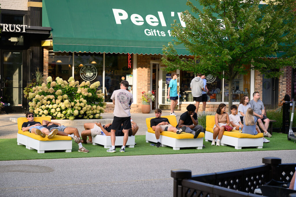 People sit and lounge on yellow and white outdoor seats in front of a shop with a green awning that reads "Peel 'n' Stick Gifts for All." Discover Midland as some chat while others relax, soaking in the welcoming atmosphere.