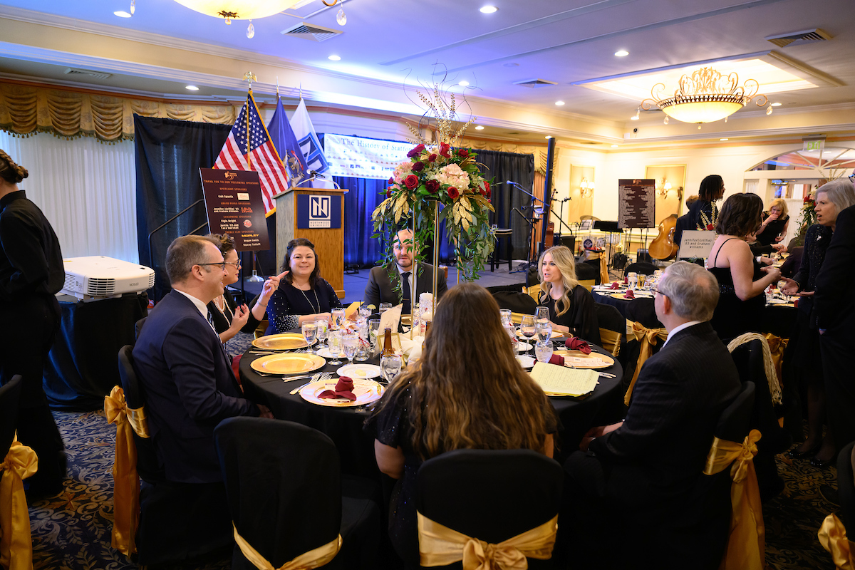 Guests in formal attire gather around a decorated banquet table at the Stafford Dinner, with a stage and elegant floral centerpiece enhancing the atmosphere in the background.