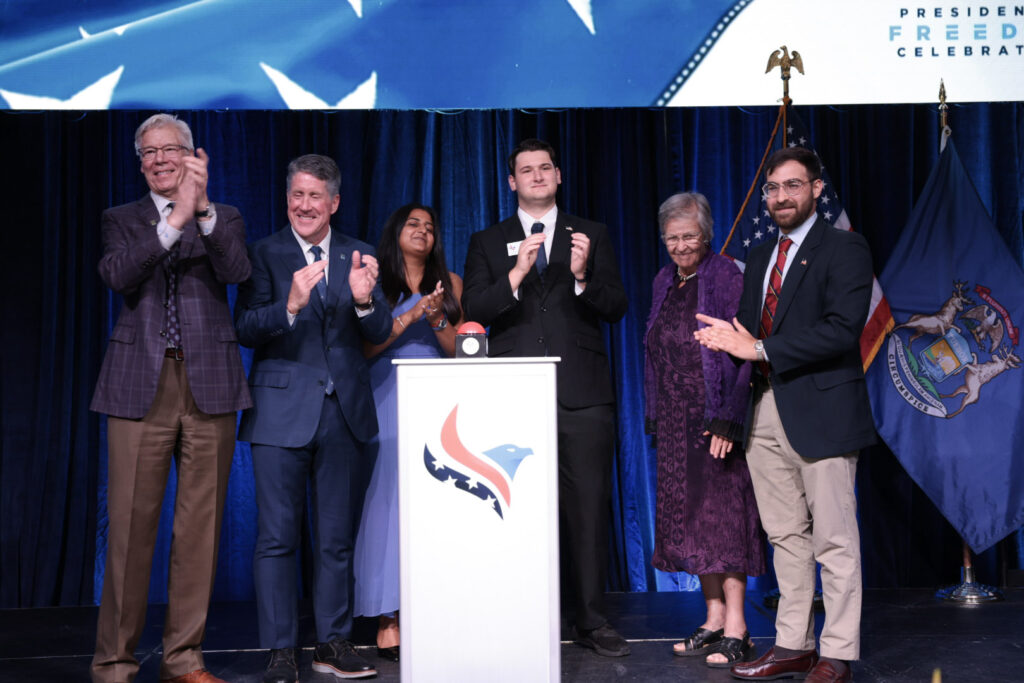 Six people in formal attire stand on a stage clapping, with a podium in front and flags in the background. The setting appears to be an official or celebratory Northwood event, possibly linked to the $100M "When We Are Free" campaign.