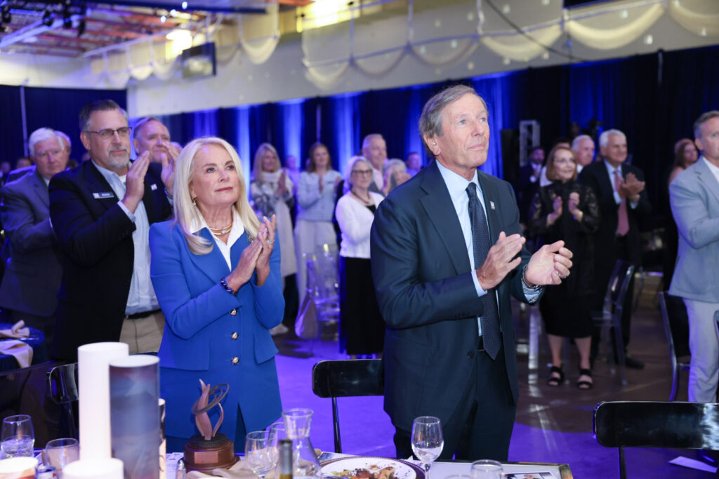 A group of people dressed in formal attire stand and applaud at a Northwood indoor event with blue lighting and decorated tables, celebrating the $100M When We Are Free Campaign.