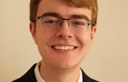 Young man with short light brown hair and glasses, wearing a black suit, white shirt, striped tie, and two pins, smiling in front of a plain light-colored wall—capturing a confident student view on nuclear safety.