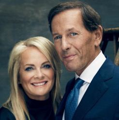 A woman with long blonde hair and a man in a blue suit and tie pose together, smiling, in front of a dark background.