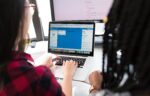 Two people are working together at a desk, looking at a laptop screen displaying a project management or collaboration tool.