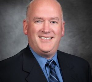 A man in a suit and tie smiles at the camera against a dark, professional studio background.