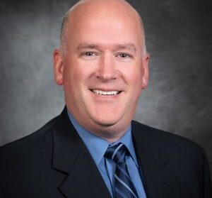 A man in a suit and tie smiles at the camera against a dark, professional studio background.