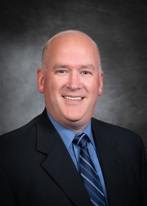 A man in a suit and tie smiles at the camera against a dark, professional studio background.