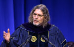 A person in academic regalia speaks at a podium with two microphones against a blue curtain background.