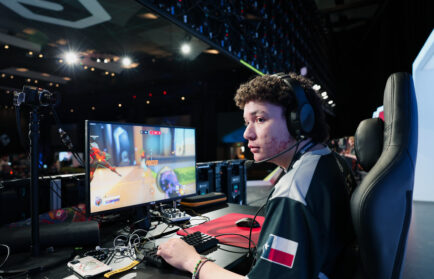 A gamer wearing a headset sits at a desk playing a first-person shooter game on a monitor in a large, well-lit venue with gaming equipment and audience in the background.
