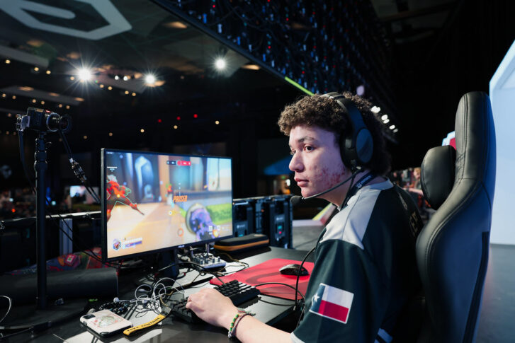 A gamer wearing a headset sits at a desk playing a first-person shooter game on a monitor in a large, well-lit venue with gaming equipment and audience in the background.
