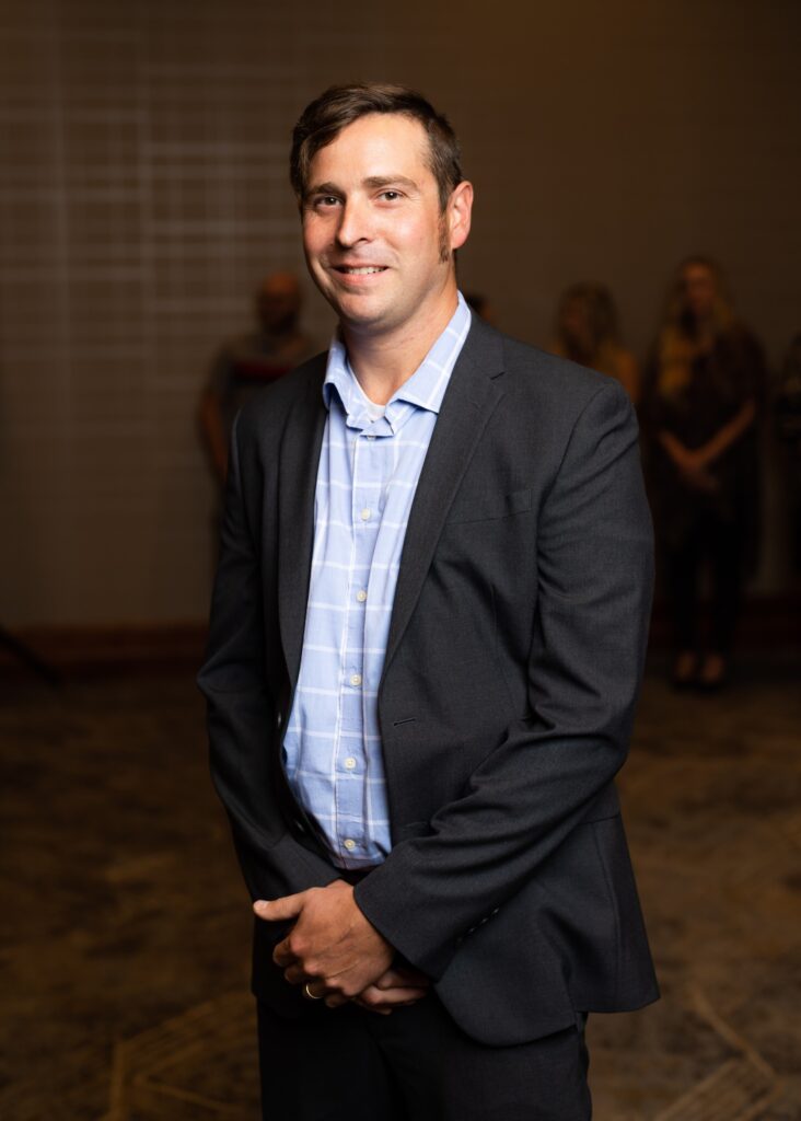 A man in a dark suit and light blue checked shirt stands indoors, facing the camera, with several people blurred in the background.