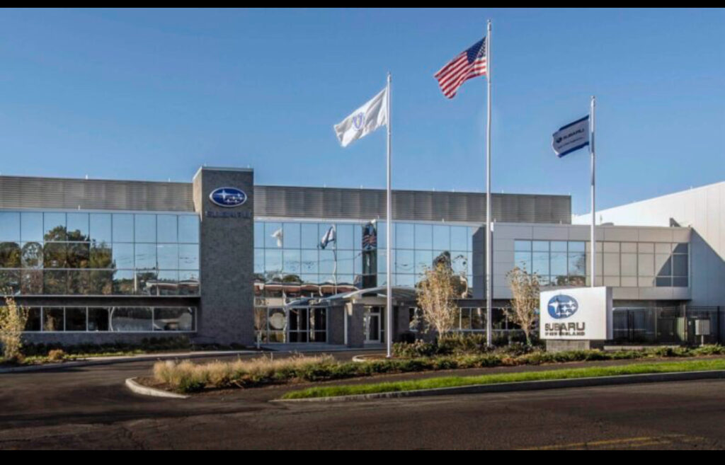 A Subaru building with three flagpoles out front, including the American flag, and a Subaru sign near the entrance. The building has large windows and a clear sky above.