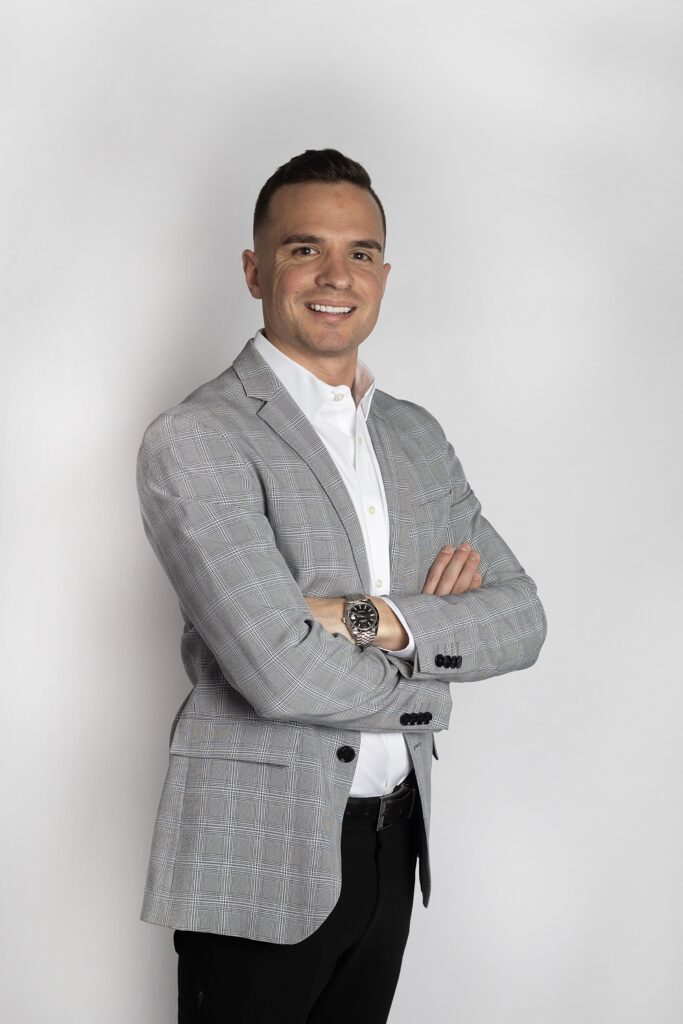 Outstanding Alumni: Man in a light gray plaid blazer and white shirt, standing with arms crossed and smiling in front of a plain white background.