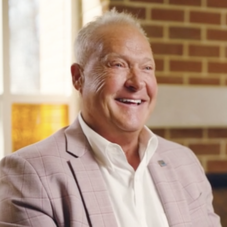 A man with short light hair in a light-colored blazer and white shirt smiles while seated indoors near a window and brick wall at the Center for Automotive and Mobility Studies.