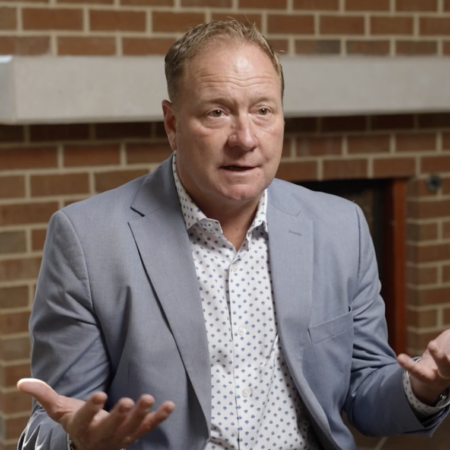 A man in a light blue blazer and patterned shirt gestures with open hands while seated indoors in front of a brick wall, representing the Center for Automotive and Mobility Studies.