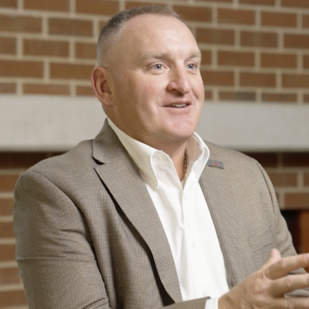 A man in a tan blazer and white shirt, representing the Center for Automotive and Mobility Studies, sits in front of a brick wall, gesturing with his hands while speaking.