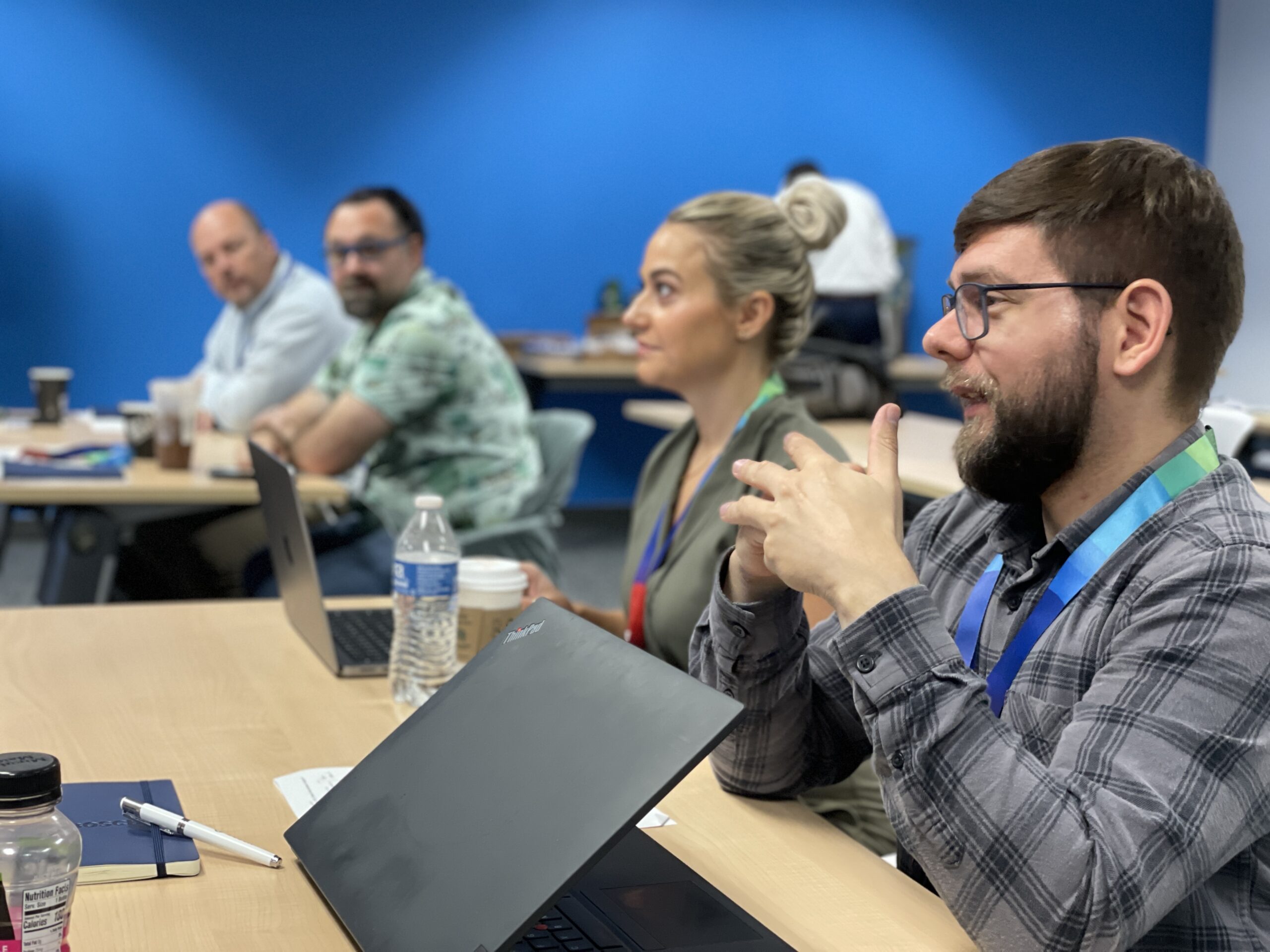 Four people sit at a conference table with laptops and notebooks, engaged in discussion about INFUSION 2025-26 in a brightly lit room with a blue wall.