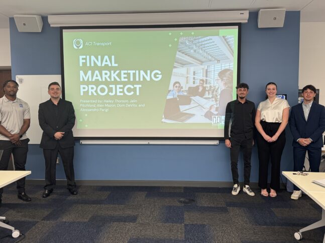 Five DeVos students stand in front of a screen displaying a slide titled "Final Marketing Project" for AC Transport, showcasing the real-world impact of their business fieldwork in a conference room.