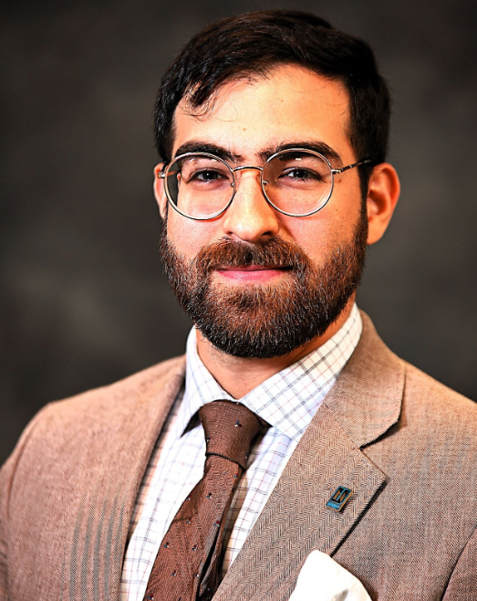 A man with dark hair and a beard wearing glasses, a light brown suit, checkered shirt, and brown tie poses for a professional portrait against a dark background.