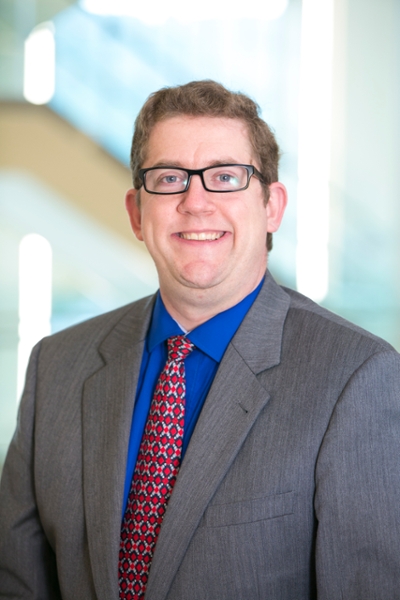 A man with short brown hair and glasses, wearing a gray suit, blue shirt, and patterned red tie, stands indoors with a blurred background.