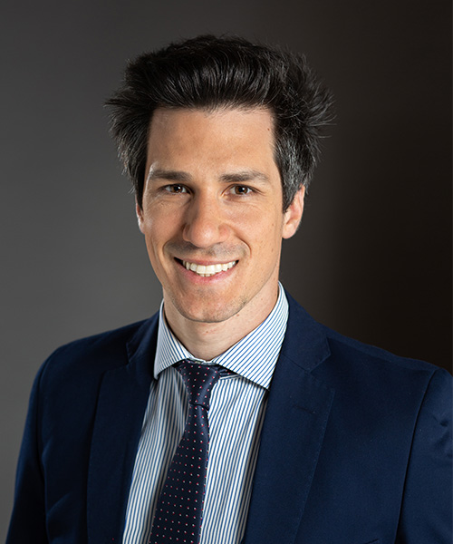 A man in a navy suit, striped shirt, and polka dot tie smiles at the camera against a plain dark background.