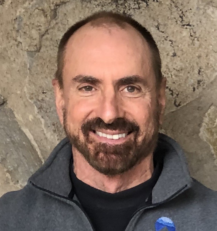 A middle-aged man with short hair and a beard smiles at the camera, wearing a gray zip-up jacket, standing in front of a stone wall.