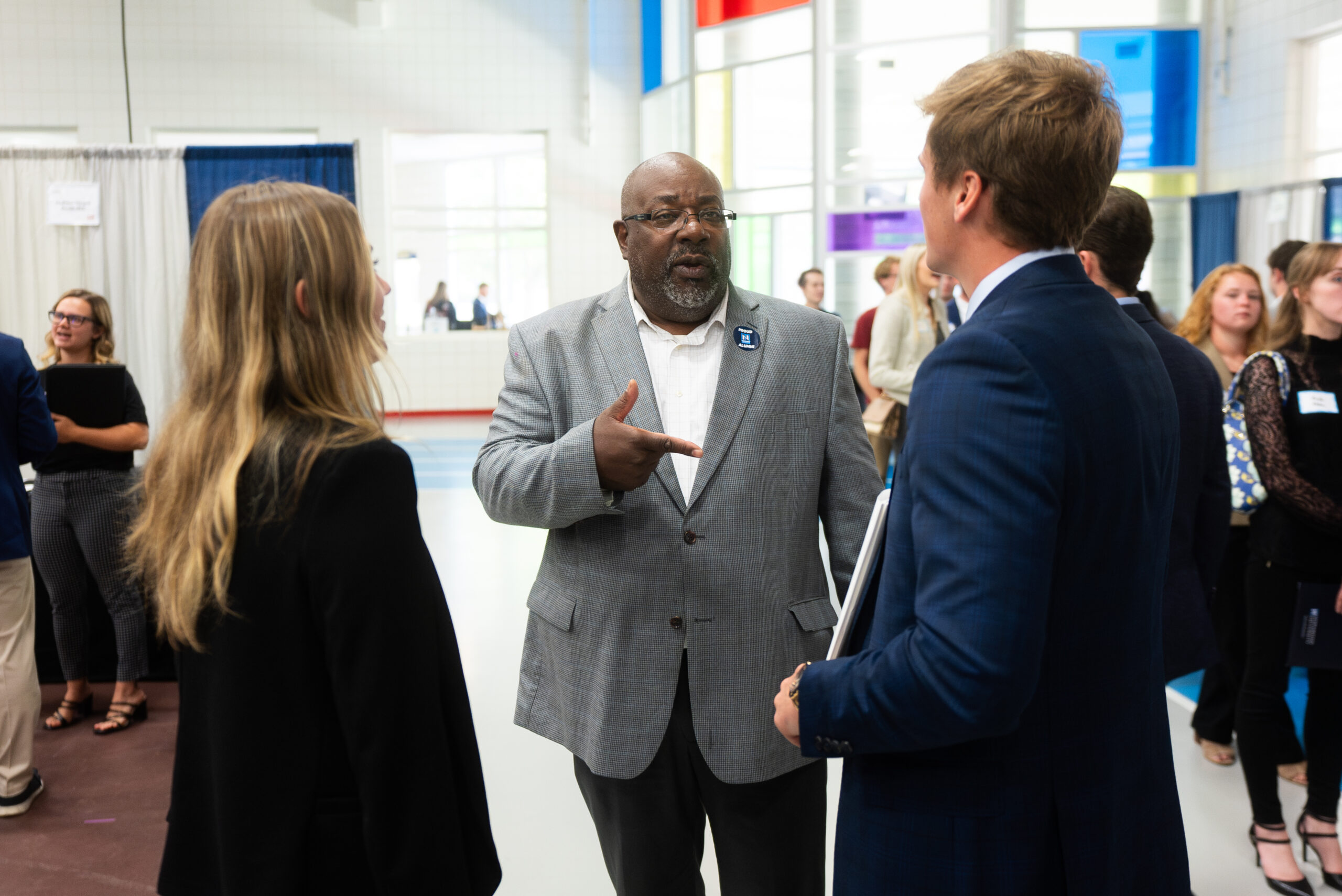 A man in a gray blazer speaks and gestures to two people in business attire at an indoor networking event, with others present in the background.