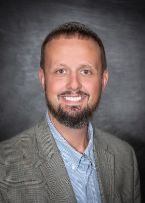 A man with short brown hair and a trimmed beard, wearing a plaid blazer and a blue collared shirt, smiles in front of a dark gray studio background.