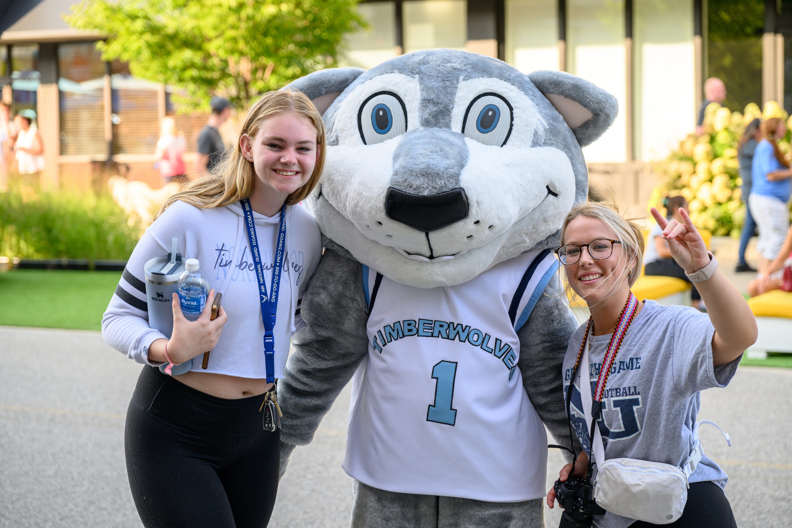 Two young women pose with a Timberwolves mascot outdoors during Go MAD March; one holds a camera, the other a water bottle, while the mascot sports a jersey at Campus to Main Street on Aug. 22.