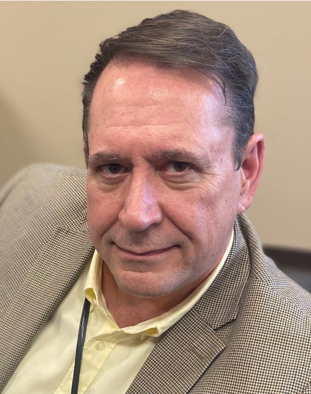 A middle-aged man wearing a light-colored dress shirt and checked blazer sits indoors, looking directly at the camera.