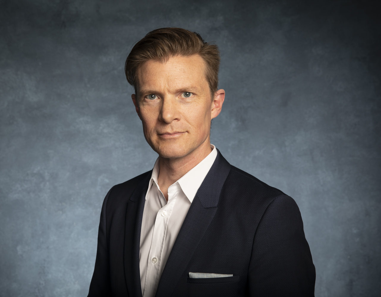 A man in a dark suit and white shirt poses against a blue-gray textured background, looking directly at the camera with a neutral expression.