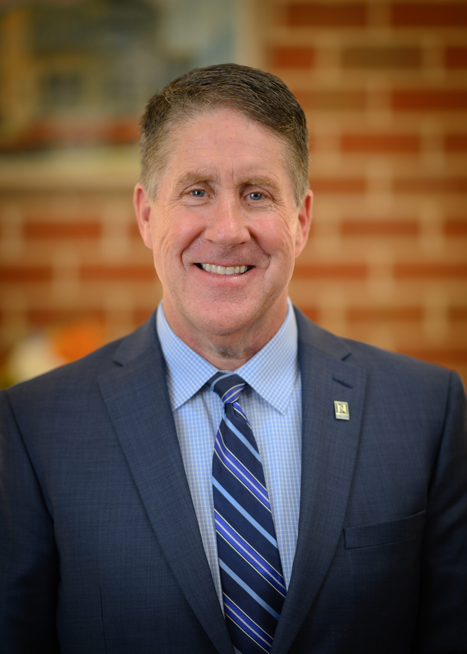 A middle-aged man in a blue suit, striped tie, and light blue shirt stands smiling in front of a brick wall background.