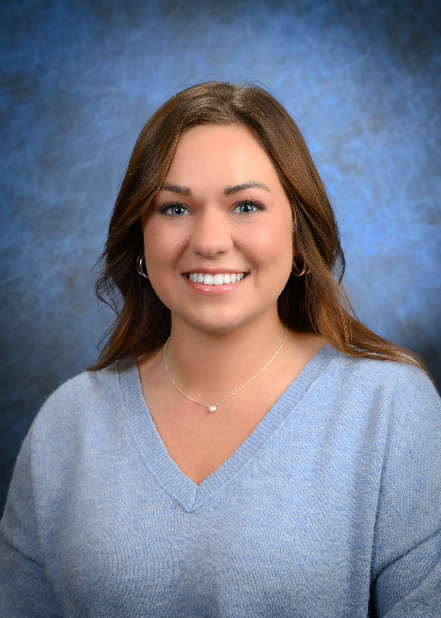 A woman with long brown hair and blue eyes smiles at the camera, wearing a light blue sweater and a necklace, with a blue mottled background.
