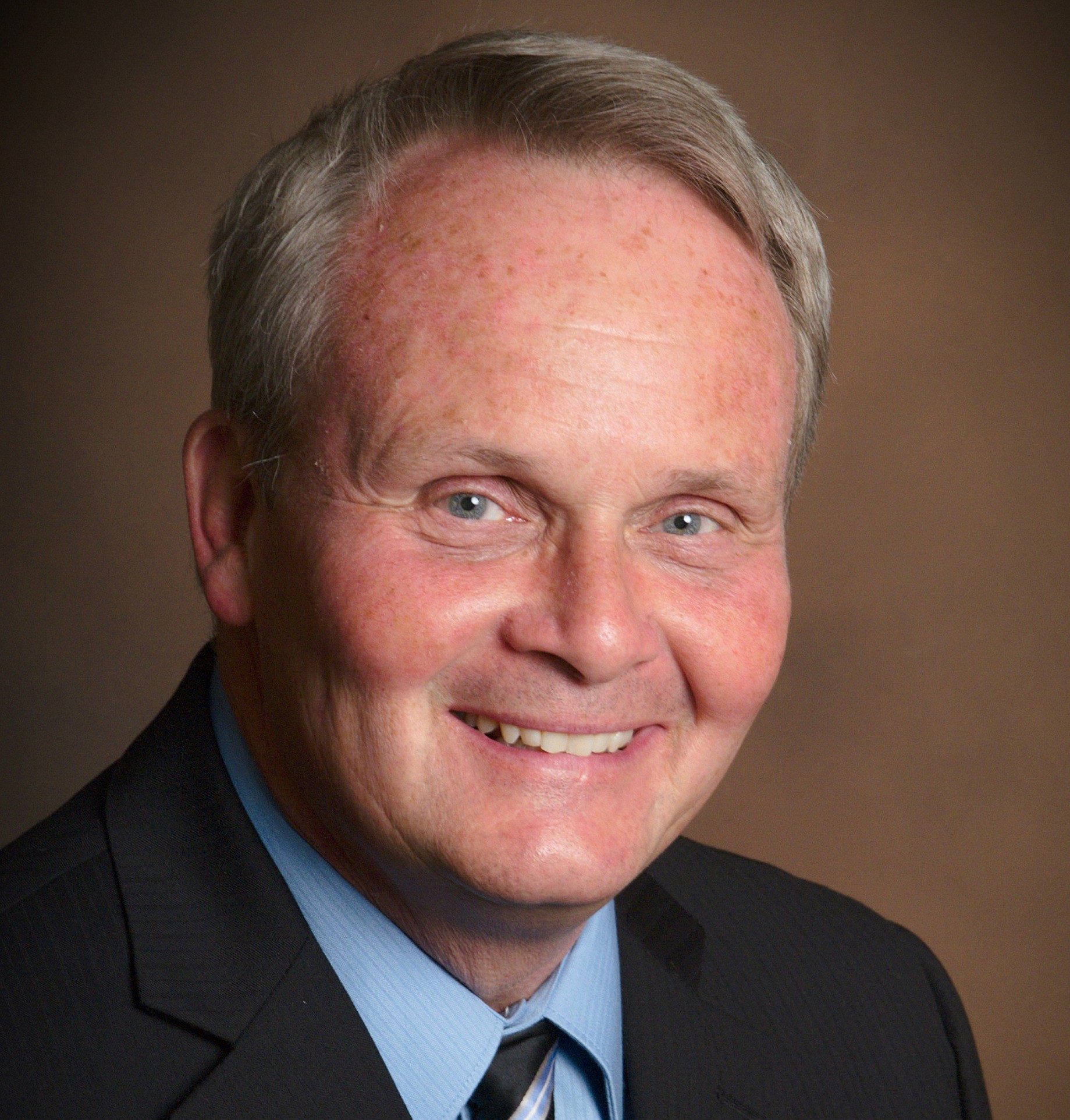A middle-aged man with short gray hair smiles, wearing a dark suit jacket, blue shirt, and striped tie against a brown background.