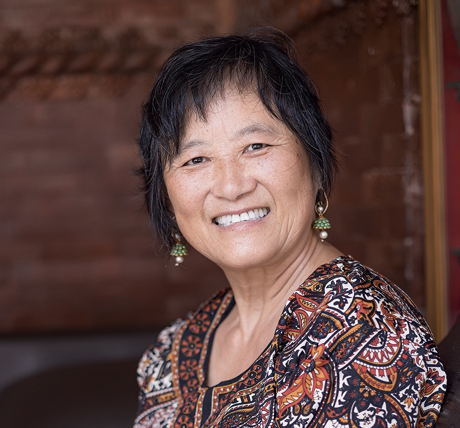 An older woman with short black hair smiles at the camera, wearing a patterned top and green earrings, with a blurred brown background.