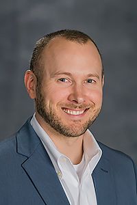 A man in a blue suit jacket and white shirt smiles at the camera against a plain, dark background.