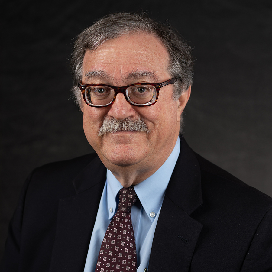 A middle-aged man with gray hair, glasses, and a mustache is wearing a dark suit, blue shirt, and patterned tie, posed against a dark background.