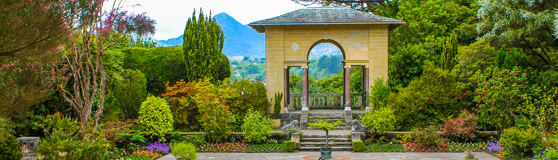 A stone archway stands in a lush, landscaped garden, reminiscent of the Emerald Isle’s verdant countryside, with vibrant plants, blooming flowers, and mountain scenery in the background.