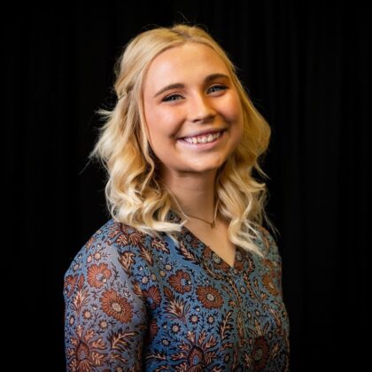 A woman with blonde, wavy hair smiles at the camera. She is wearing a blue floral patterned blouse and is posed in front of a black background.