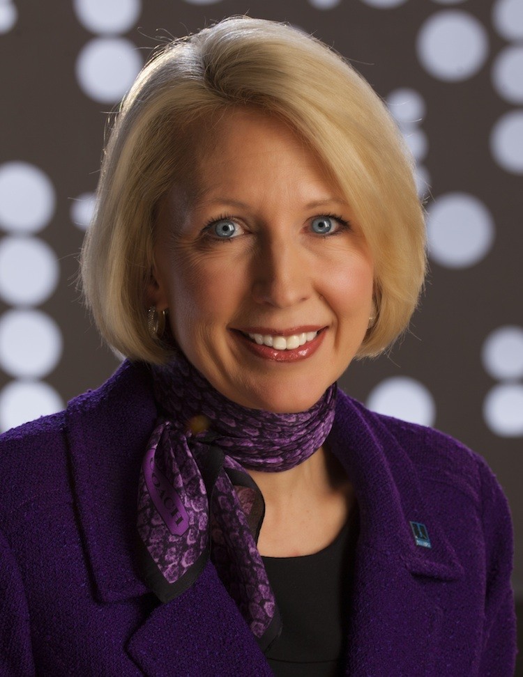 A woman with short blonde hair, wearing a purple blazer and matching scarf, smiles warmly in front of large white circular lights.
