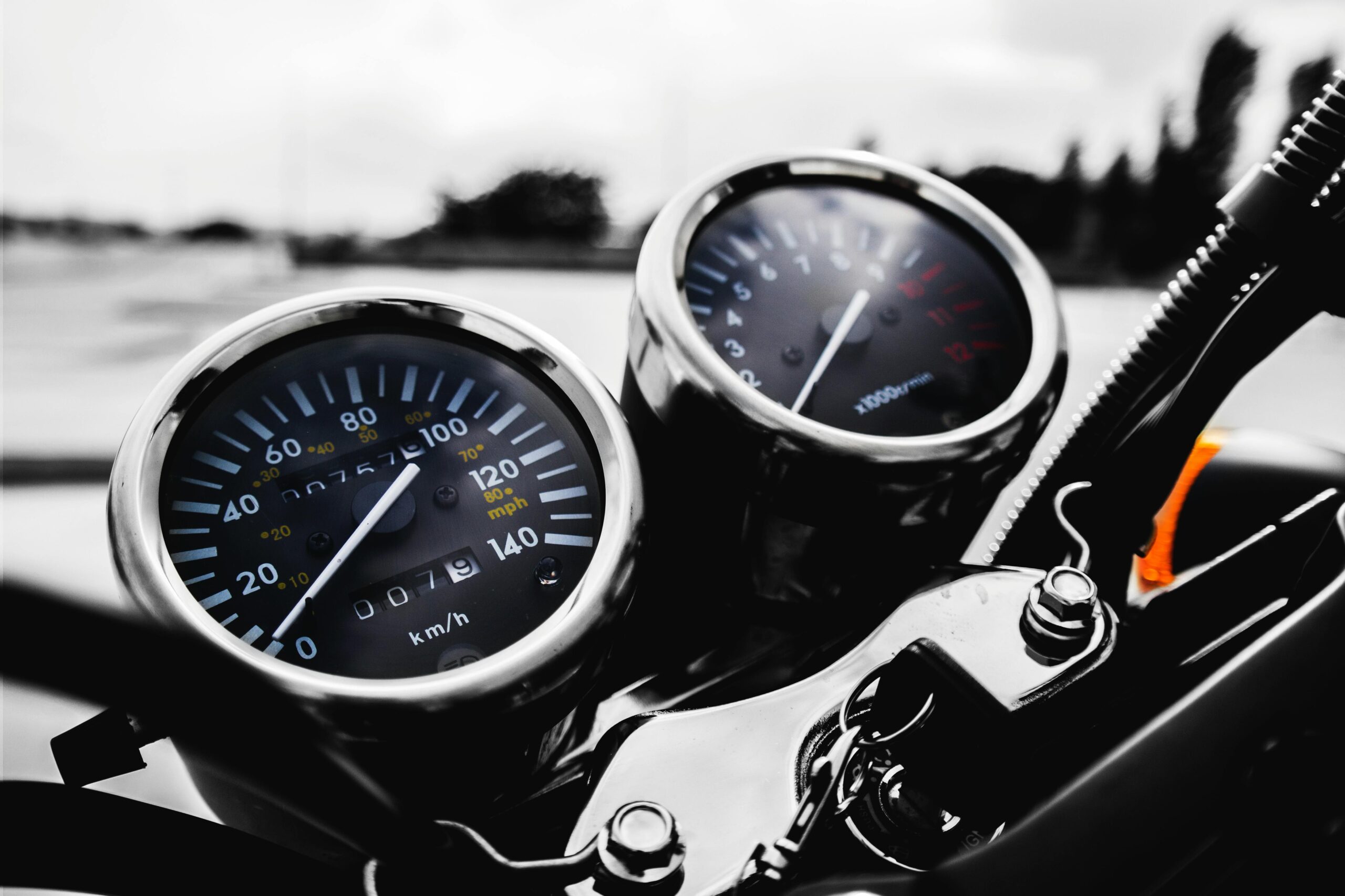 Close-up of a motorcycle dashboard showing a speedometer in kilometers per hour and a tachometer with blurred outdoor background, perfect for those interested in Automotive Management.